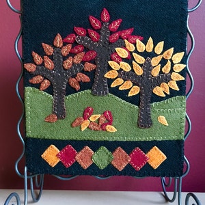 May include: A black felt wall hanging with a fall theme. Three trees with brown trunks and colorful leaves in shades of red, orange, and yellow are stitched onto a green hill. The bottom border has a row of diamond shapes.