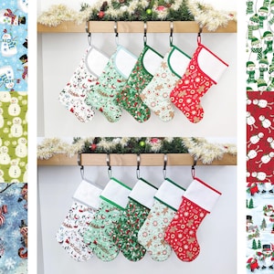 May include: Six Christmas stockings with different snowman designs hang on a wooden shelf. The stockings are white with a red, green, or blue trim. The snowman designs are all different, but they all feature snowmen with hats and scarves.