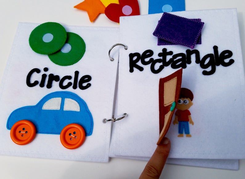 May include: A felt activity book with pages featuring shapes and objects. One page shows a blue car with orange button wheels and the word "Circle". The other page has the word "Rectangle" and a felt door with a boy.