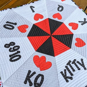 May include: A white quilted fabric play mat with a black and red center. The mat has a variety of playing card symbols and numbers printed on it, including hearts, spades, diamonds, clubs, and numbers 1, 10, and king. The mat is designed for children to play games on.