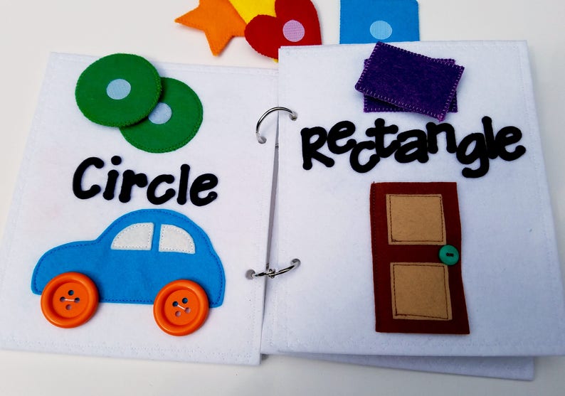May include: Open felt book with colorful shapes and words. The left page shows a green circle with a light blue center, the word "Circle", and a blue car with orange button wheels. The right page has the word "rectangle" and a brown door.