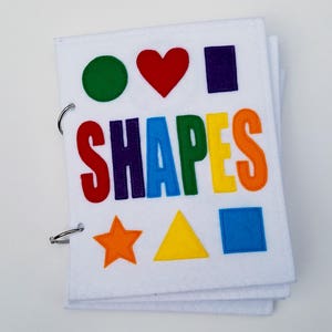 May include: A white felt book with colorful shapes and the word "SHAPES" in rainbow letters. The shapes include a green circle, red heart, purple rectangle, orange star, yellow triangle, and blue square. The book is bound with metal rings.