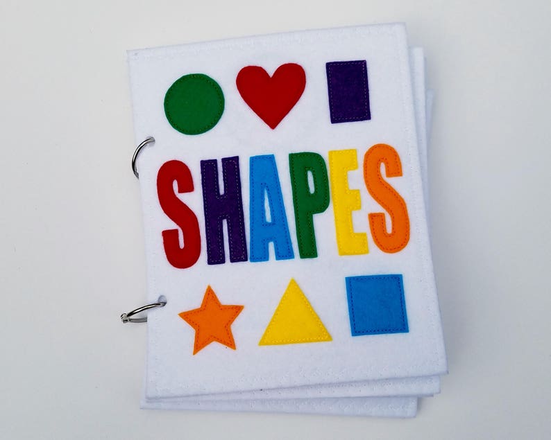 May include: A white felt book with colorful shapes and the word "SHAPES" in rainbow letters. The shapes include a green circle, red heart, purple rectangle, orange star, yellow triangle, and blue square. The book is bound with metal rings.