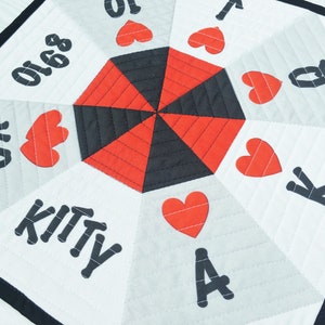 May include: A white fabric game board with a red and black center. The board has a black border and features playing card symbols, including hearts, kings, queens, aces, and numbers. The text "KITTY" is printed on the board.