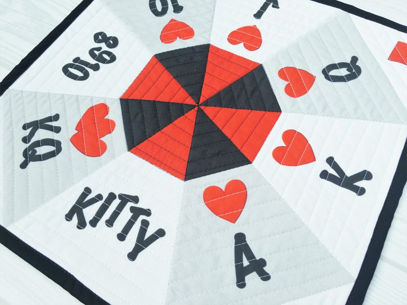 May include: A white fabric game board with a red and black center. The board has a black border and features playing card symbols, including hearts, kings, queens, aces, and numbers. The text "KITTY" is printed on the board.