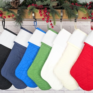 May include: A row of seven quilted Christmas stockings in various colors, including black, navy, blue, green, white, cream, and red. Each stocking has a white cuff and is hanging from a metal hook. Festive greenery and red berries are above.