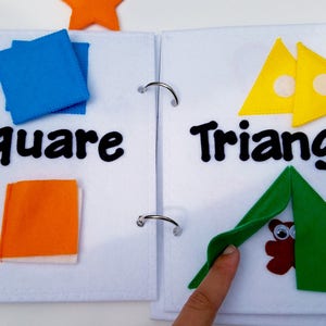 May include: A felt activity book open to two pages. One page shows blue squares and the word "Square". The other page displays yellow triangles and the word "Triangle". A green triangle is folded over a brown owl.