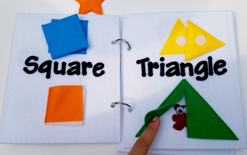 May include: A felt activity book open to two pages. One page shows blue squares and the word "Square". The other page displays yellow triangles and the word "Triangle". A green triangle is folded over a brown owl.