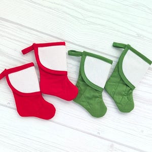 May include: Two Christmas stockings, one red and one green, with white felt trim and a white canvas panel for personalization. The stockings are on a white wood background.