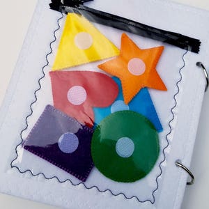 May include: A white felt activity book with a clear pocket containing colorful felt shapes: yellow triangle, orange star, red heart, blue triangle, green circle, and purple square. Each shape has a white velcro dot. The book has a black zipper and metal rings.