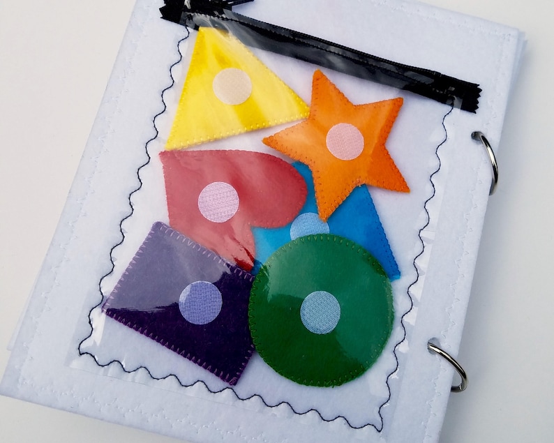 May include: A white felt activity book with a clear pocket containing colorful felt shapes: yellow triangle, orange star, red heart, blue triangle, green circle, and purple square. Each shape has a white velcro dot. The book has a black zipper and metal rings.