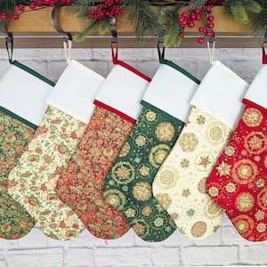 Könnte beinhalten: Sechs Weihnachtsstrümpfe mit verschiedenen floralen und geometrischen Mustern in Rot, Grün und Creme. Jeder Strumpf hat eine weiße Manschette mit farbigem Besatz und eine Aufhängeschlaufe. Die Strümpfe werden auf einem Holzregal mit Grünzeug ausgestellt.