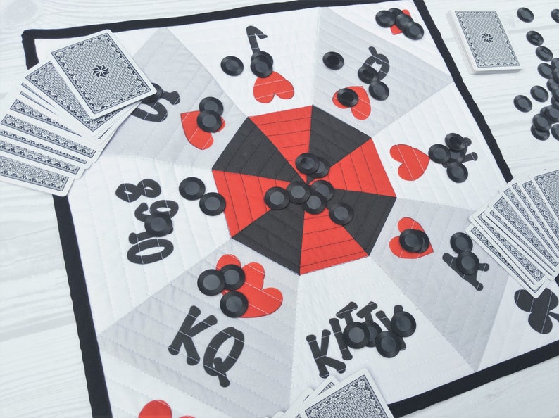 May include: A white fabric game board with a red and black octagon in the center. The board has black felt circles and hearts around the octagon. The board has the words "King", "Queen", "Jack", and "Ace" written on it. There are also stacks of playing cards on the board.