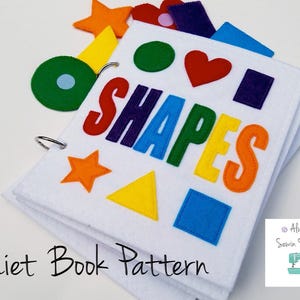 May include: A white felt quiet book with colorful felt shapes and the word "SHAPES" in large, multi-colored letters. The book is bound with a metal ring and includes the text "Quiet Book Pattern".