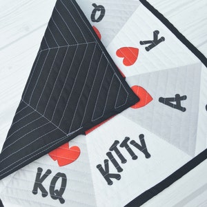 May include: A white and gray quilted fabric square with black trim. The fabric features a design of playing cards with red hearts and black suits. The text "Kitty" is printed in black on the fabric.