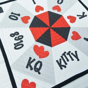 May include: A white fabric game board with a black and red hexagon in the center. The board has black numbers and letters, including "Kitty", "KQ", "A", "K", "10", "8", and "Q". There are also red hearts around the hexagon.