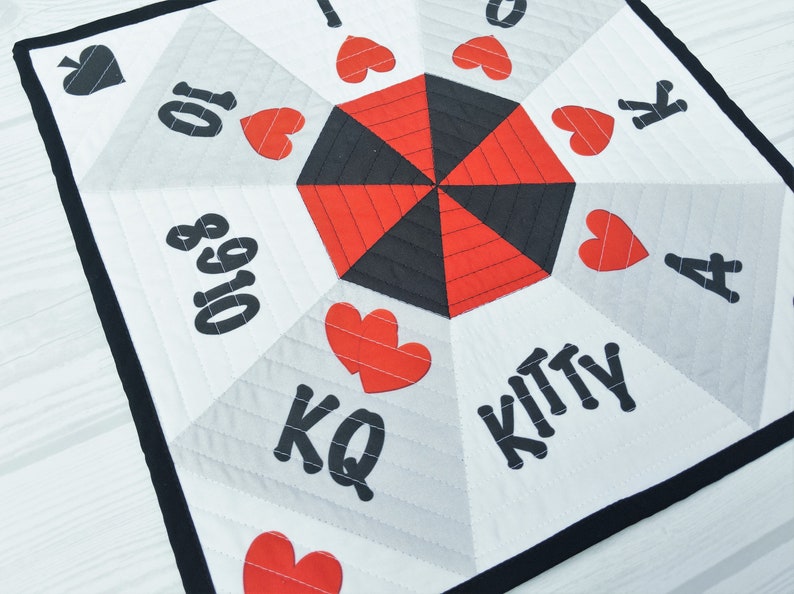 May include: A white fabric game board with a black and red hexagon in the center. The board has black numbers and letters, including "Kitty", "KQ", "A", "K", "10", "8", and "Q". There are also red hearts around the hexagon.