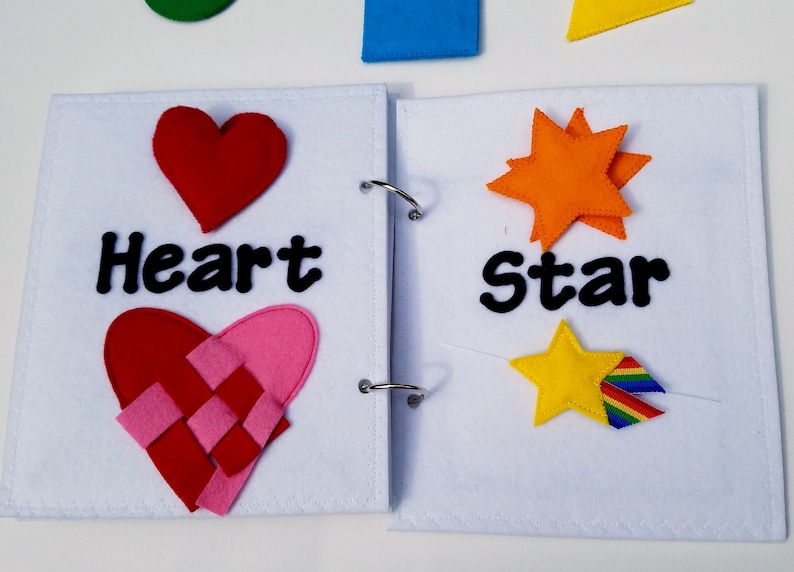 May include: A felt book with white pages and metal rings. The left page has a red heart and the word "Heart". The right page has an orange star and the word "Star". Additional felt shapes are visible.