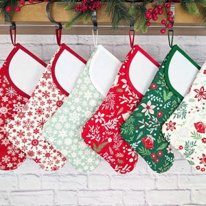 May include: Six Christmas stockings with various festive patterns, including snowflakes, floral designs, and botanical prints. Each stocking has a white interior and a contrasting trim in red or green. They hang from a decorative metal rod.