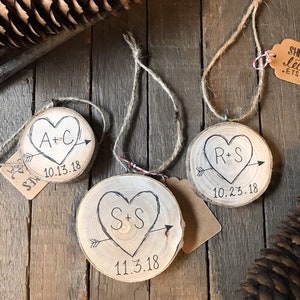 May include: Three wooden ornaments with heart-shaped designs and initials. Each ornament has a date and an arrow through the heart. The ornaments are hanging from twine.