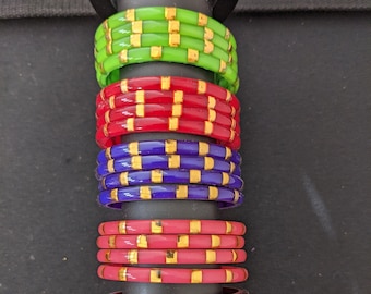 May include: A stack of colorful glass bangles with gold accents. The bangles are arranged in a vertical stack on a black stand. The bangles are green, red, blue, pink, brown, and yellow.