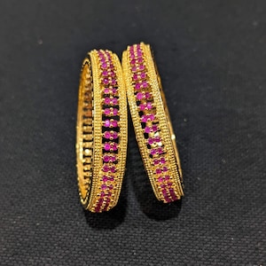 May include: Two gold bangles with a row of pink gemstones around the circumference.