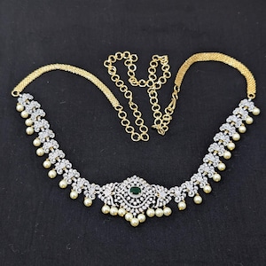 May include: A gold-toned necklace with a central emerald-colored stone and surrounding clear stones. The necklace features a series of small, round, white pearls and a chain extension for adjustable sizing. The background is black.