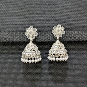 May include: A pair of silver-toned jhumka earrings. Each earring features a floral design at the top, a bell-shaped body with mirrored accents, and a fringe of small white beads. The earrings are displayed against a dark gray background.