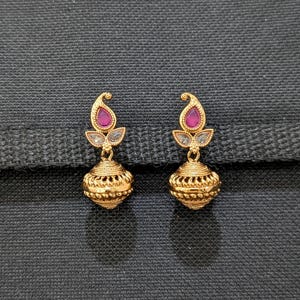 May include: A pair of gold-colored earrings. Each earring features a teardrop-shaped, magenta-colored stone at the top, with a small, clear stone below. The lower portion of the earrings is a round, textured design.