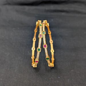 May include: Two gold bangles with a repeating pattern of clear stones and alternating red and green square accents. The bangles are set against a dark background, highlighting their intricate design and sparkle.
