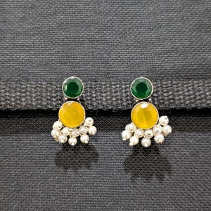 May include: A pair of silver-toned stud earrings. Each earring features a small, round, dark green gemstone at the top, a larger yellow gemstone in the middle, and a cluster of small, white, pearl-like beads at the bottom. The earrings are set against a dark gray background.