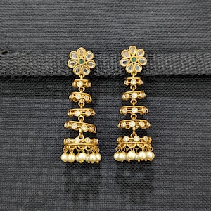 May include: A pair of gold-toned chandelier earrings. Each earring features a floral stud with a green center stone, followed by tiered circular designs with clear stones, and a pearl-beaded bottom. The earrings are displayed against a dark background.