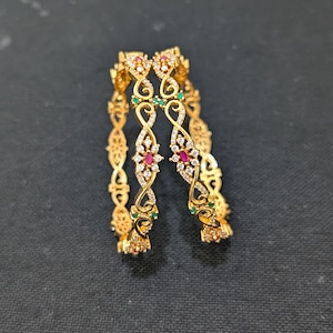 Op de afbeelding: Twee gouden armbanden met ingewikkelde bloemen- en krulwerkontwerpen. De armbanden zijn versierd met kleine rode, groene en heldere edelstenen. De achtergrond is effen zwart.