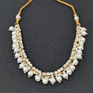 May include: A gold necklace with a delicate design. The necklace features multiple strands of small, round, white pearls. The pearls are interspersed with small, clear, round stones. The necklace is displayed against a black background.
