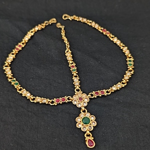 Może przedstawiać: Złoty nakrycie głowy z kwiatowym wzorem. Nakrycie głowy posiada centralny kwiat z zielonym kamieniem szlachetnym i czerwoną zawieszką w kształcie łzy. Ozdobione jest czerwonymi, zielonymi i przezroczystymi kamieniami szlachetnymi.