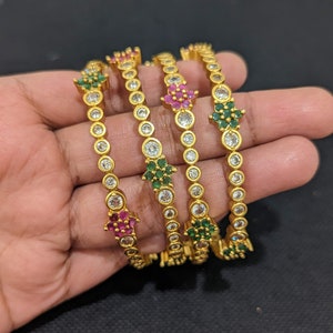 May include: Four gold bangles with white stones and colorful accents. The bangles are stacked on top of each other and feature a floral design.
