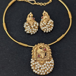 May include: A gold-toned necklace and earring set featuring a peacock motif with small white pearls and tiny red stones. The necklace has a rigid circular band with textured segments and a central pendant, while the earrings match the pendant design.