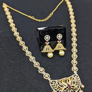 May include: A gold-toned jewelry set featuring a necklace and matching earrings. The necklace has a pendant with a peacock design and is adorned with clear gemstones. The earrings have a bell shape with a pearl drop. The set is displayed on a black surface.