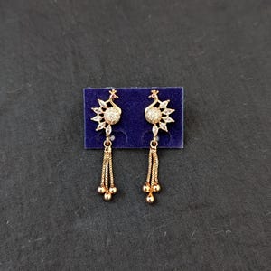 Könnte beinhalten: Ein Paar goldfarbene Pfau-Ohrringe. Jeder Ohrring hat ein Pfau-Design mit einem kristallartigen Körper und einem Fächer aus Kristallen. Von dem Pfau hängen drei Stränge einer goldfarbenen Kette, die in drei kleinen goldfarbenen Perlen enden.