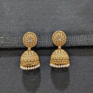 May include: A pair of gold-colored jhumka earrings. Each earring features a circular top with a central stone and intricate filigree detailing. The bell-shaped bottom is also adorned with filigree and small white bead accents.