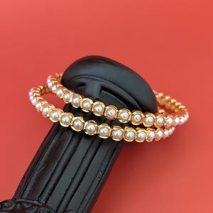 Op de afbeelding: Twee goudkleurige armbanden met witte parelaccenten rusten op een zwarte decoratieve zuil tegen een rode achtergrond. De armbanden zijn cirkelvormig, waarbij elke parel dicht bij elkaar is geplaatst. De zuil heeft een textuur.