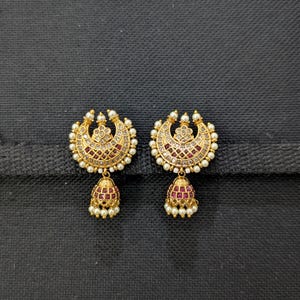 Puede incluir: Un par de pendientes dorados. Cada pendiente presenta una parte superior en forma de media luna, adornada con pequeñas perlas blancas y piedras preciosas rojas. Un colgante en forma de campana, también con piedras preciosas rojas y perlas, cuelga debajo.