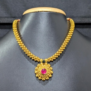 May include: A gold necklace with a beaded chain and a pendant. The pendant has a central red gemstone and a floral design. The necklace is displayed on a black mannequin.