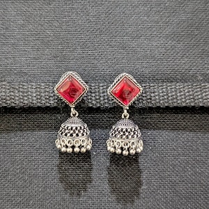 May include: A pair of silver-toned jhumka earrings. Each earring features a red, square-cut gemstone at the top, with a detailed, bell-shaped bottom. The earrings have a textured design and small dangling beads.