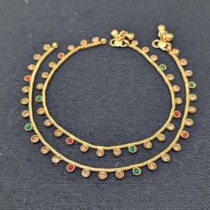 May include: A pair of gold-colored anklets with a delicate design. Each anklet features small, round, colored gemstones in red, green, and clear. The anklets are displayed on a dark background.