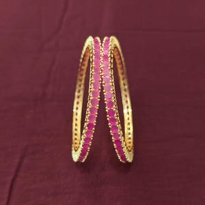 May include: Two gold-toned bangles featuring a continuous row of round, vibrant pink stones set along the outer edge. The metal bands have a textured, ridged finish on the sides, providing a metallic contrast to the bright, circular pink gemstones.