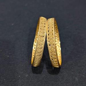 Puede incluir: Dos pulseras doradas con intrincados diseños en espiral. Las pulseras son de color dorado brillante y pulido y parecen estar hechas de metal. El diseño presenta una serie de motivos circulares conectados, creando un aspecto detallado y ornamentado.