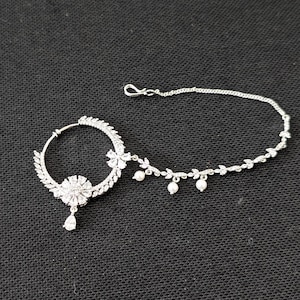 May include: A silver-toned nose ring featuring a circular hoop adorned with clear crystals and a central floral motif. A teardrop-shaped crystal hangs from the flower. A delicate chain with leaf-shaped crystal accents and small white pearls attaches.