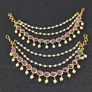 May include: A pair of gold-toned ear chains featuring multiple strands of small white pearls. The lower sections are adorned with circular motifs containing pink marquise-cut stones, central green round stones, and small white accent stones with pearl drops.