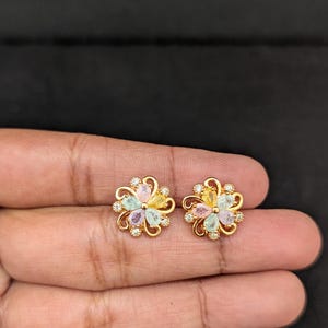 May include: A pair of gold-toned flower-shaped stud earrings. Each earring features a central cluster of pastel-colored gemstones, surrounded by delicate gold petals and small clear crystals. The earrings are displayed on a hand against a dark background.